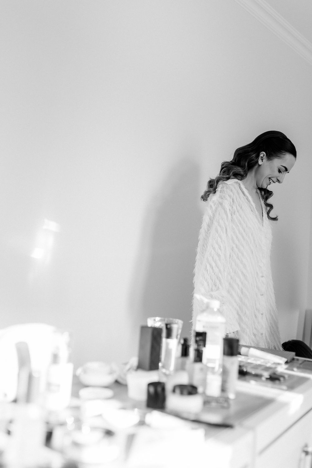Black and white photo of a smiling bride in a white robe standing near a vanity table with makeup products.