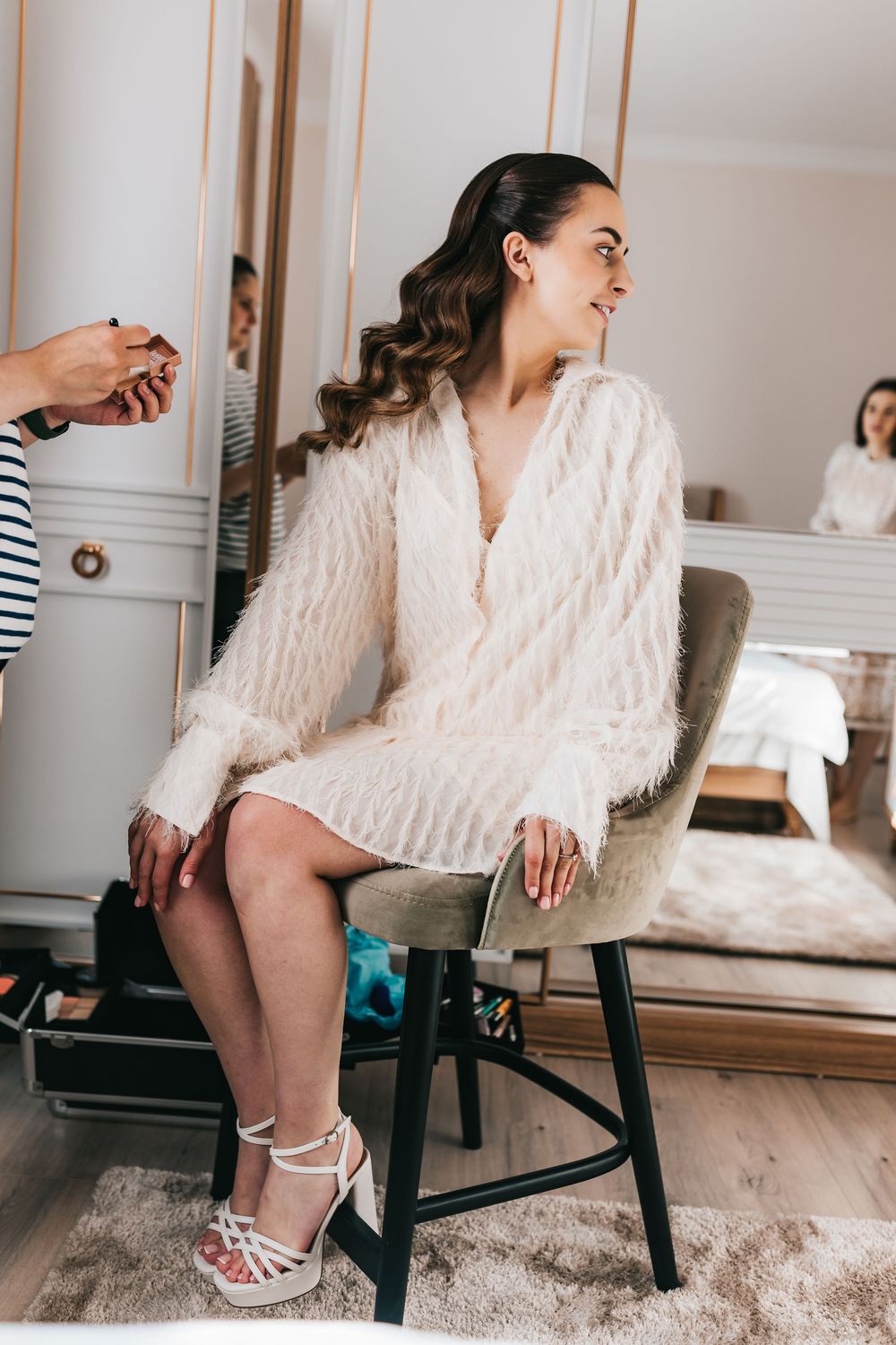 Bride in white fluffy robe and heels sits in chair getting ready, with mirror reflection in background.
