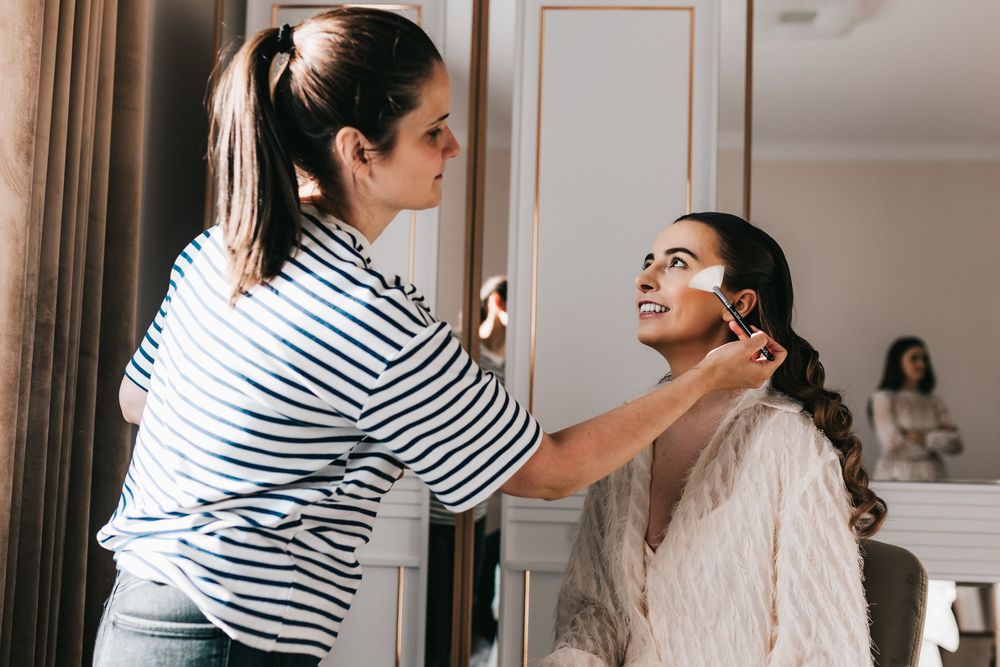 Makeup artist applying cosmetics to smiling bride in white robe during wedding preparations.