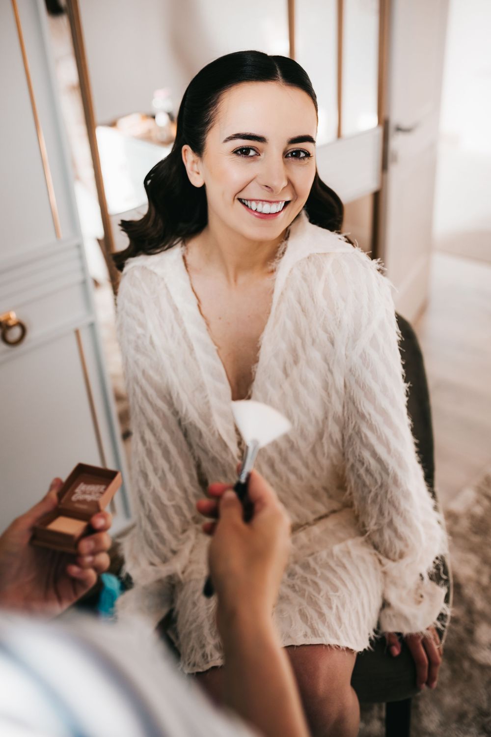 Smiling brunette bride in white fluffy robe gets makeup applied by artist before wedding ceremony.