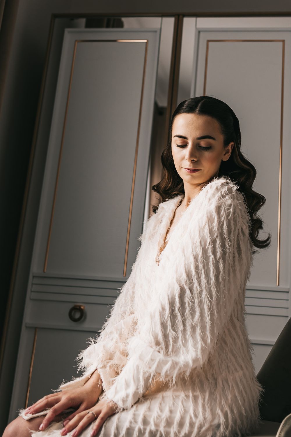 Elegant bride in white feather robe sitting gracefully before a classic wardrobe mirror.