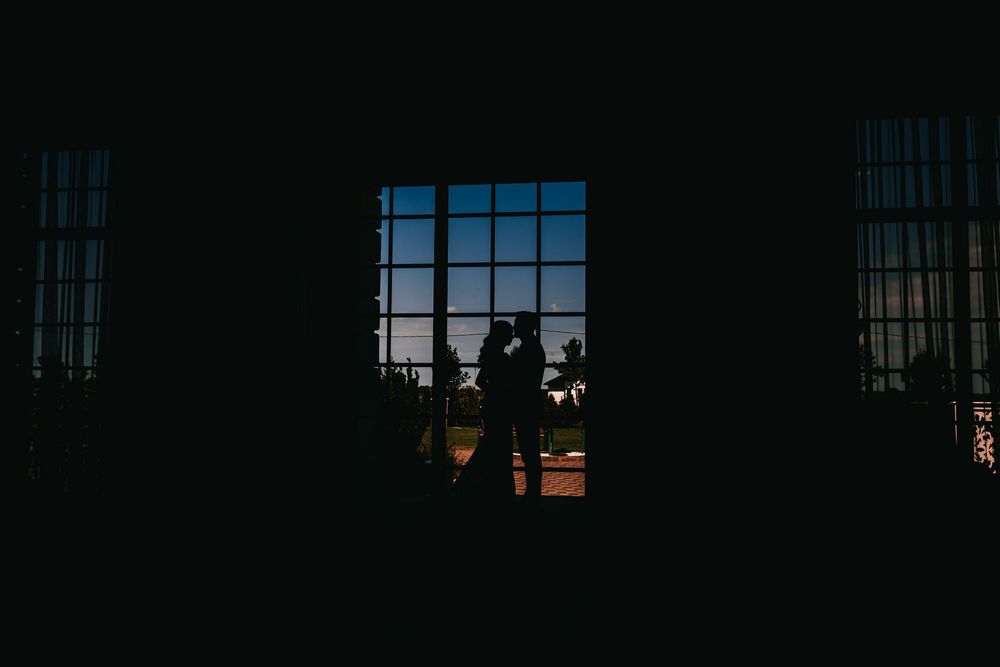Romantic silhouette of bride and groom embracing before large window at dusk.