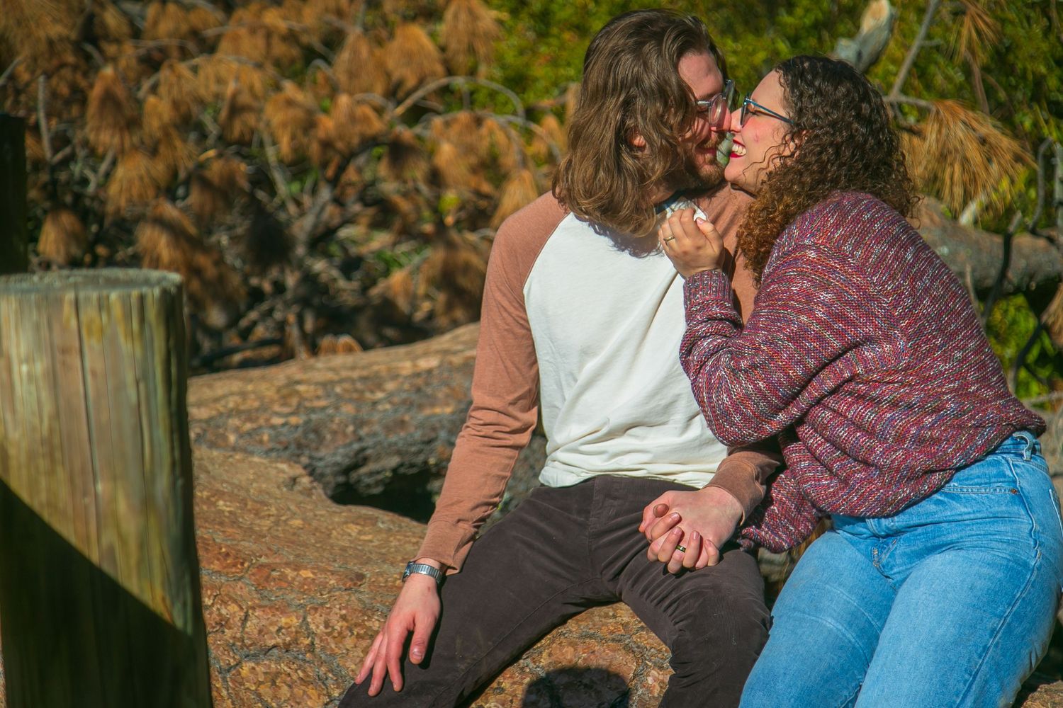 Two women share a romantic moment sitting together outdoors, surrounded by autumn foliage and wooden fence posts.