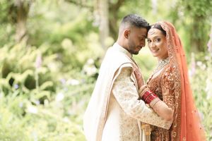South Asian bride and groom in traditional wedding attire embrace outdoors, surrounded by lush green foliage.
