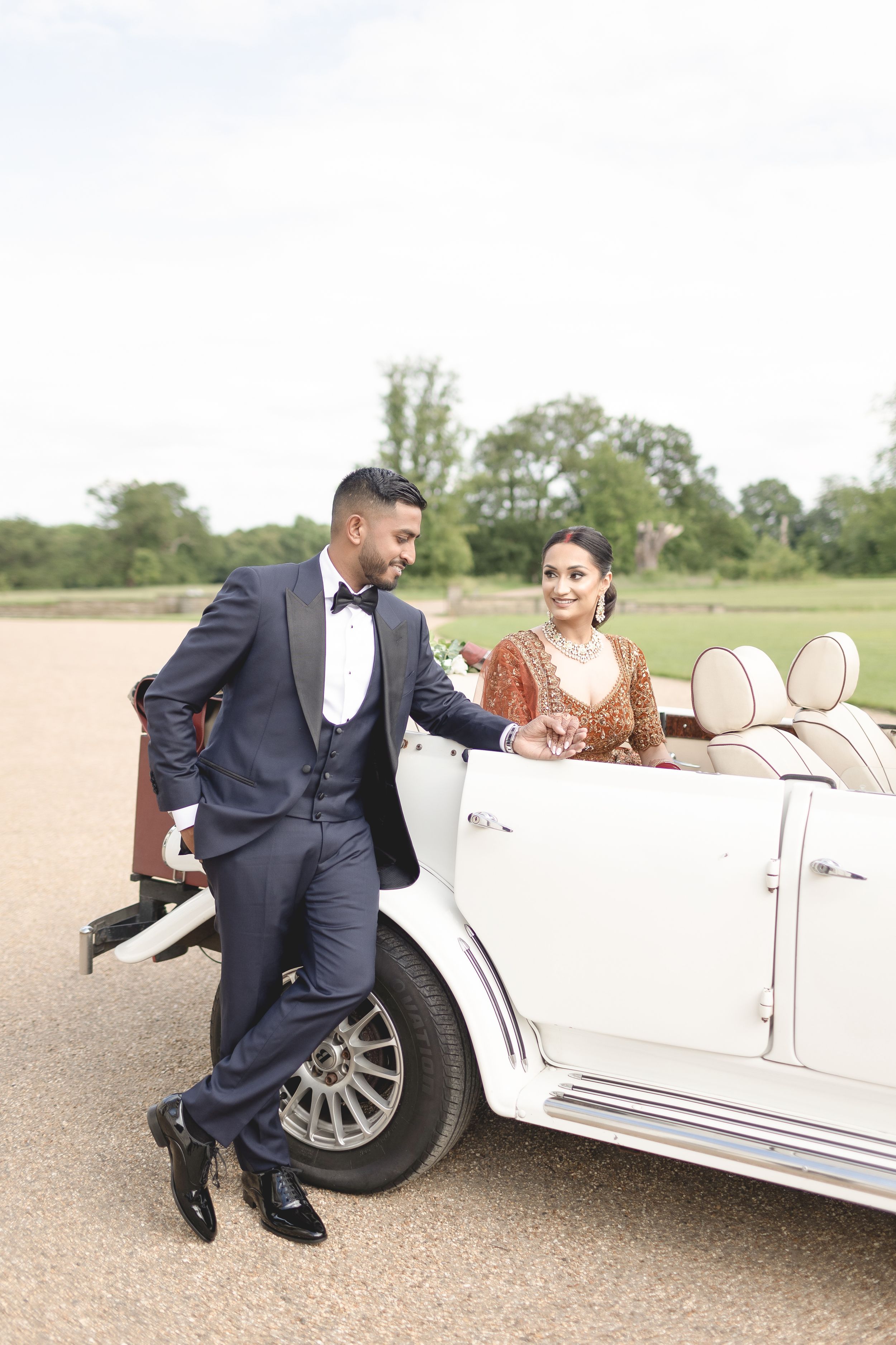 A bride and groom in elegant wedding attire pose beside a vintage white convertible car outdoors.