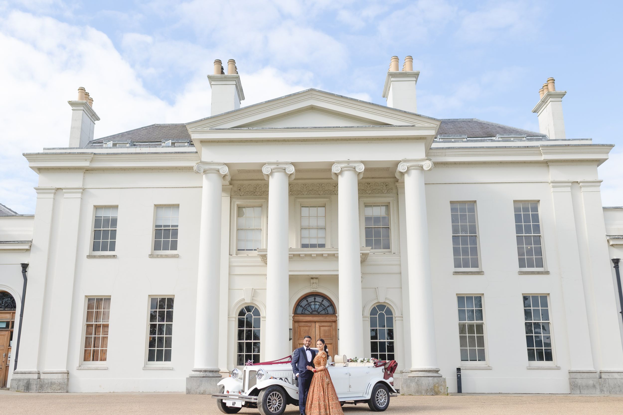 Bride and groom pose with vintage white car outside grand neoclassical mansion wedding venue with tall columns.