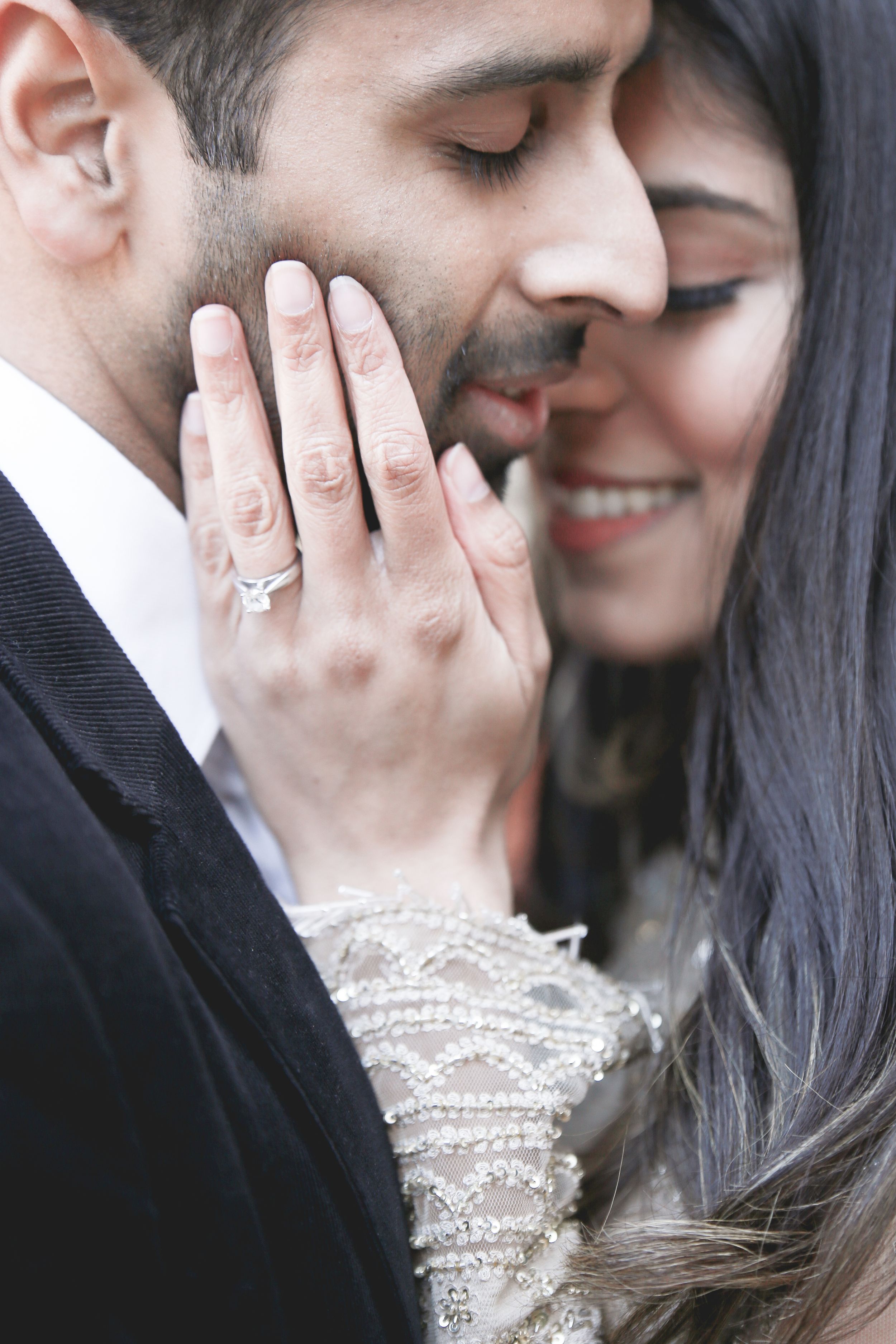 Couple in close embrace showing diamond engagement ring, woman smiling with eyes closed, man in black suit.