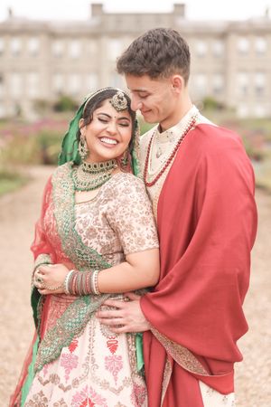 A smiling bride in green and pink Indian bridal attire embraces a groom in red shawl and cream sherwani outdoors.