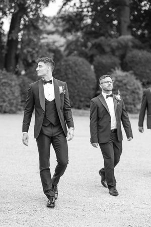 Two groomsmen in black tuxedos with bow ties and boutonnieres walking outdoors in a black and white wedding photo.