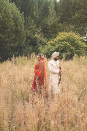 Sikh bride in red lehenga and groom in white sherwani standing in golden meadow with lush green trees backdrop.