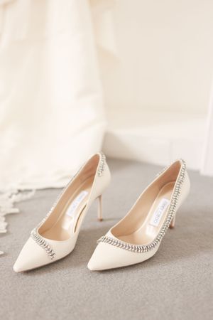Elegant ivory Jimmy Choo bridal pumps with crystal embellishments displayed on gray surface near wedding dress.