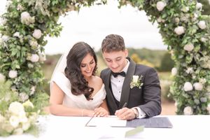 Bride and groom signing wedding register under floral arch with white roses and greenery.