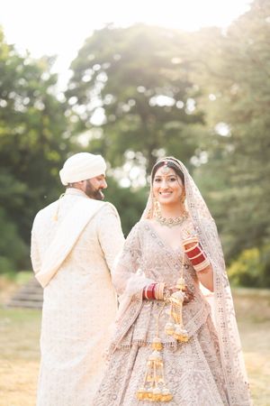 Indian bride in silver lehenga smiles at groom in white sherwani during outdoor wedding ceremony with golden sunlight.
