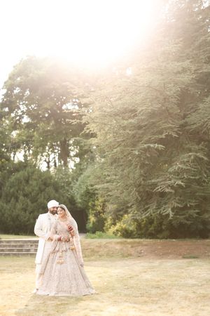 South Asian bride and groom in traditional attire pose outdoors in golden sunlight surrounded by lush green trees.