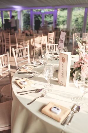 Elegant wedding reception table setting with white linens, crystal glasses, pink floral centerpiece, and table number card in bright venue.
