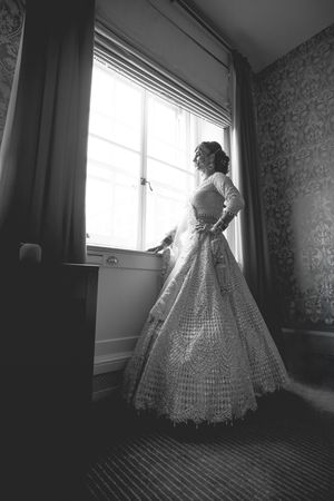 Black and white photo of a bride in an elegant ballgown standing by a window in a ornate room.