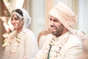 Smiling groom in white sherwani and turban during Indian wedding ceremony with bride in background.