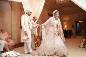 Bride and groom in traditional white Indian wedding attire exchange vows during elegant indoor ceremony with floral garlands.