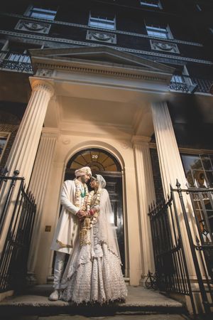 Indian bride and groom in traditional white wedding attire share a romantic moment on elegant London townhouse steps at night.