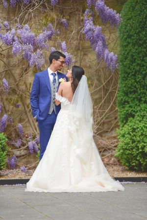 Bride and groom share a romantic moment surrounded by purple wisteria blooms at their outdoor wedding ceremony.