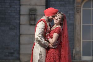 Indian couple in traditional wedding attire, bride in red lehenga and groom in cream sherwani, sharing a romantic moment.