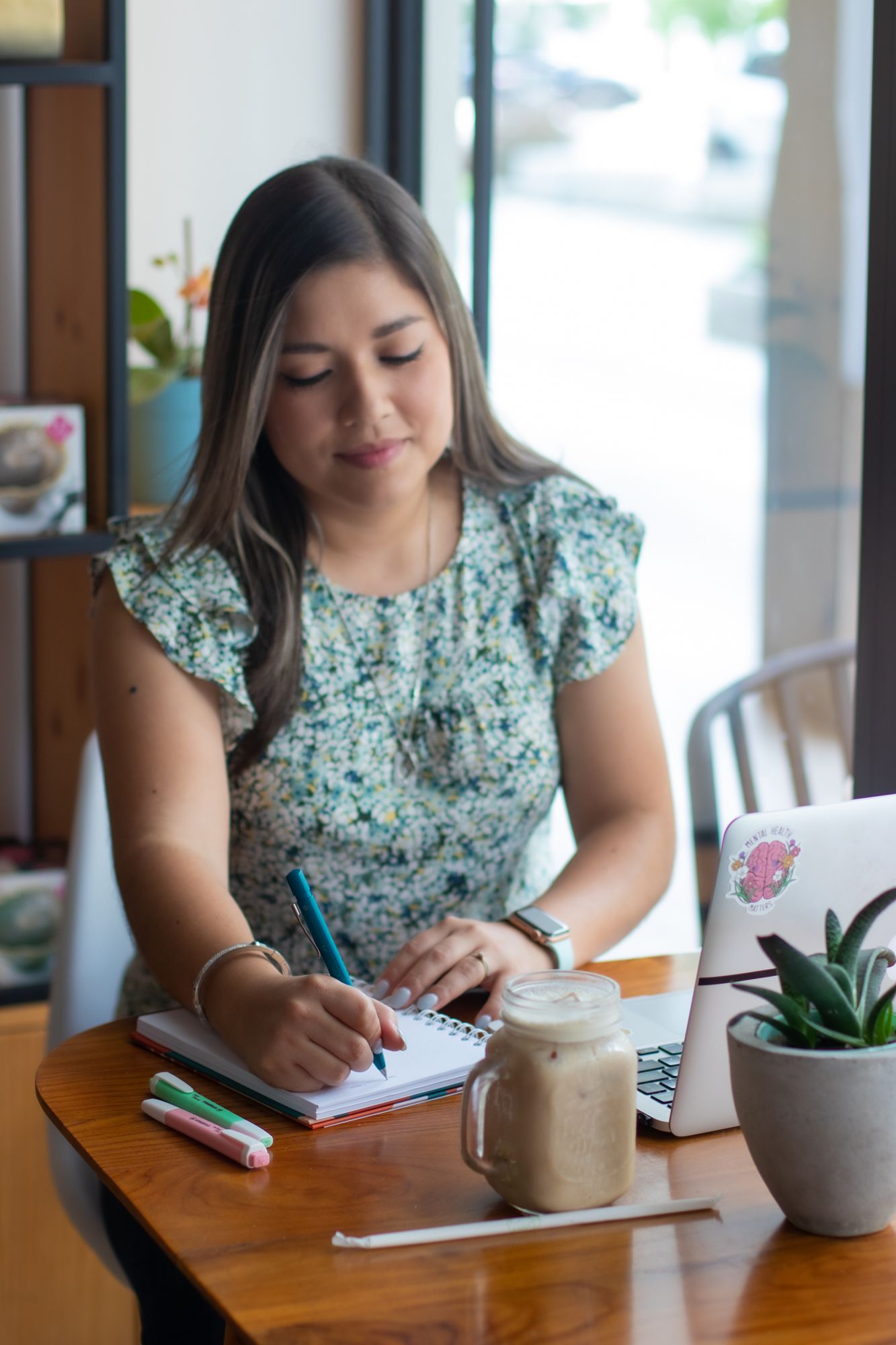 Woman in floral dress writing in notebook at cafe table with iced coffee and laptop.