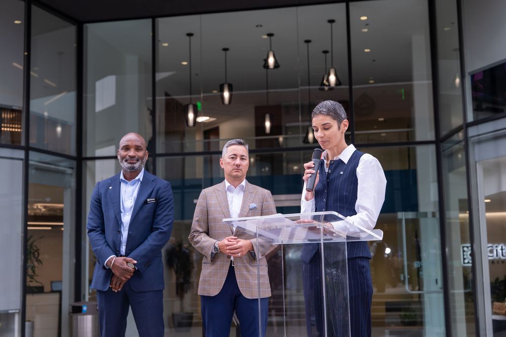 Three professionals stand at a podium outside a modern building during a formal speaking event.