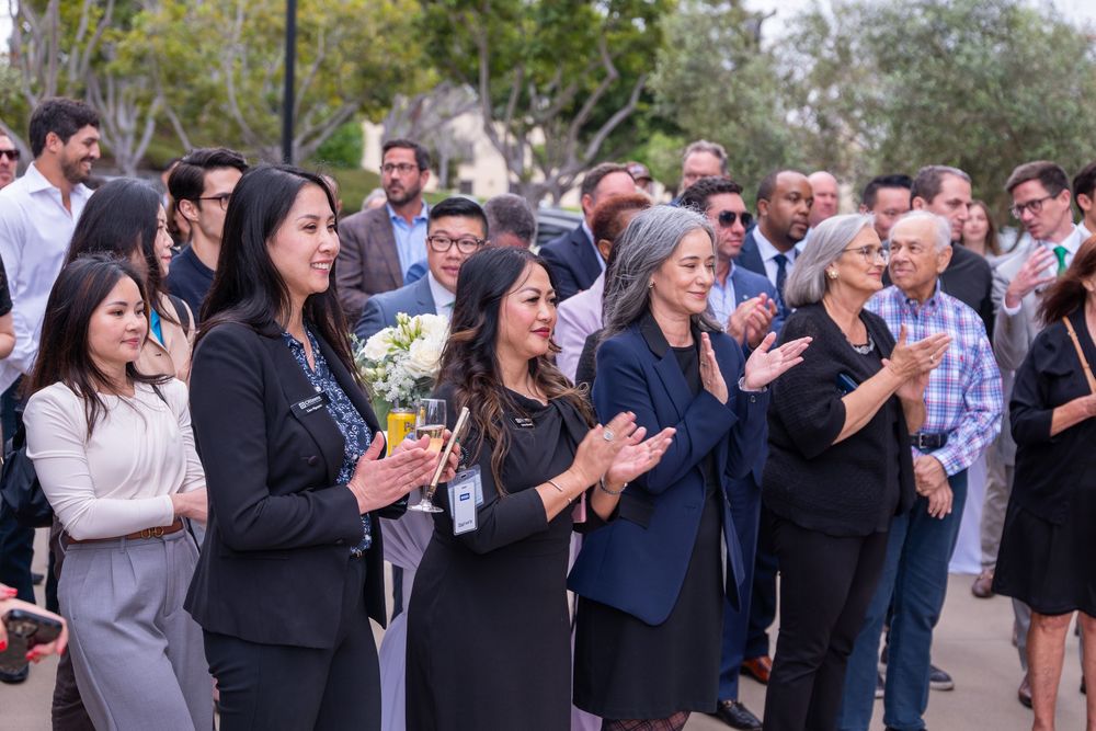 Diverse group of professionals applauding outdoors at a formal gathering, with a woman holding white flowers in front.