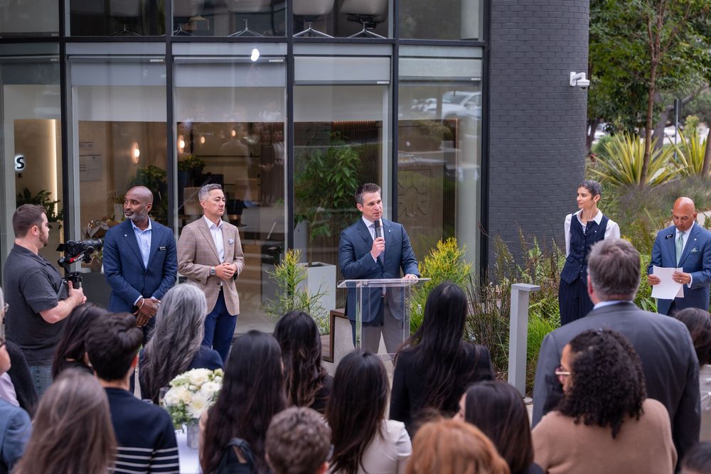 A speaker addresses a crowd at an outdoor corporate event with panelists standing near a modern glass building.