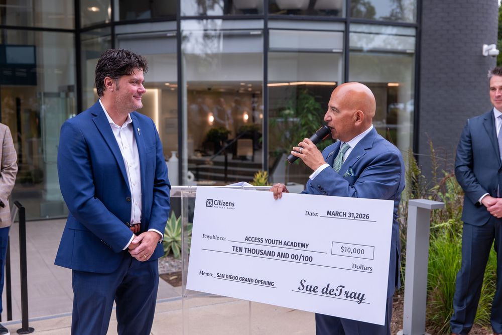 Two men smile while presenting a $10,000 ceremonial check to Access Youth Academy outdoors.