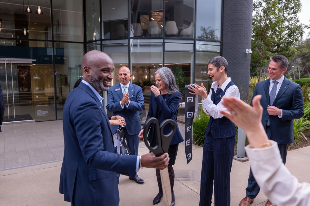 Group of professionals in business attire applauding outdoors near a modern glass building during a formal ceremony.