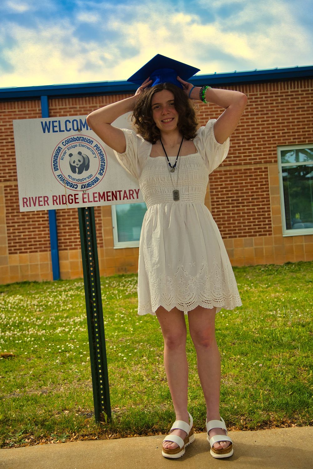 Graduate in white dress holding blue cap outside River Ridge Elementary School sign.