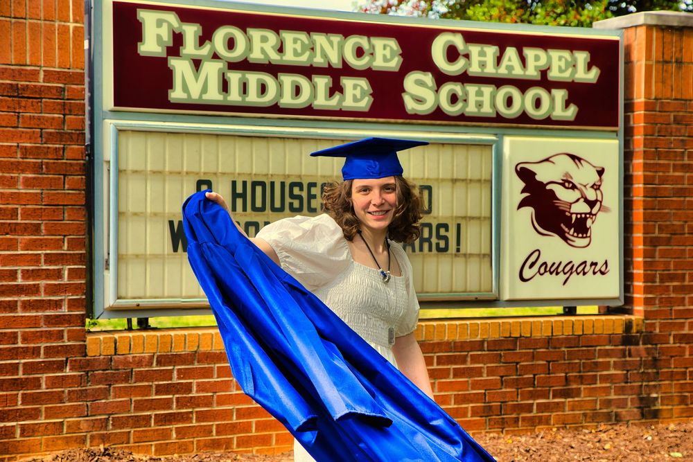 Graduate in blue cap and gown posing at Florence Chapel Middle School Cougars sign.