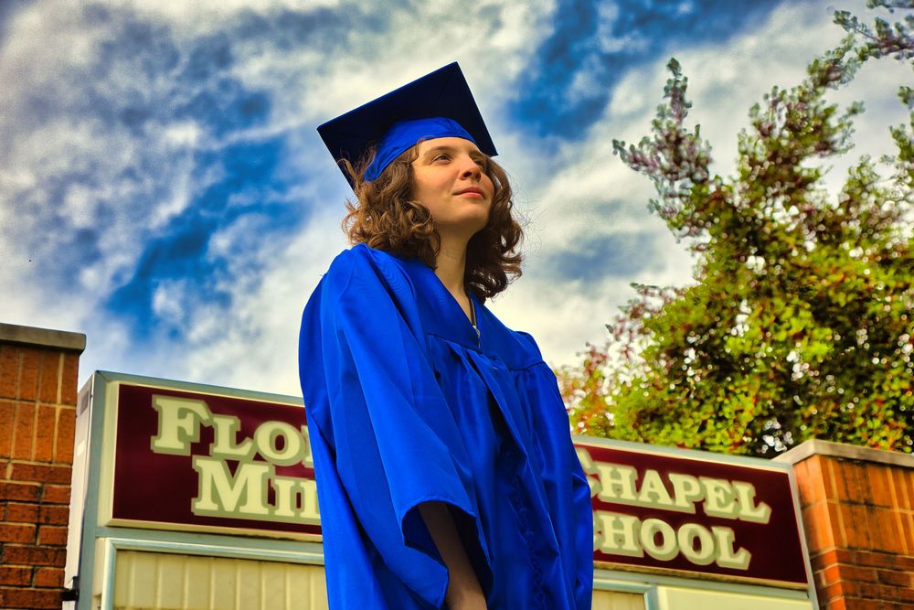 Graduate in blue cap and gown looking up at sky outside Florence Chapel Middle School.