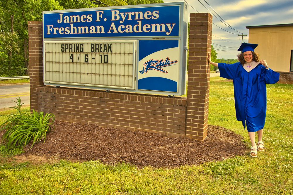 Smiling graduate in blue cap and gown posing by James F. Byrnes Freshman Academy sign in Duncan SC.