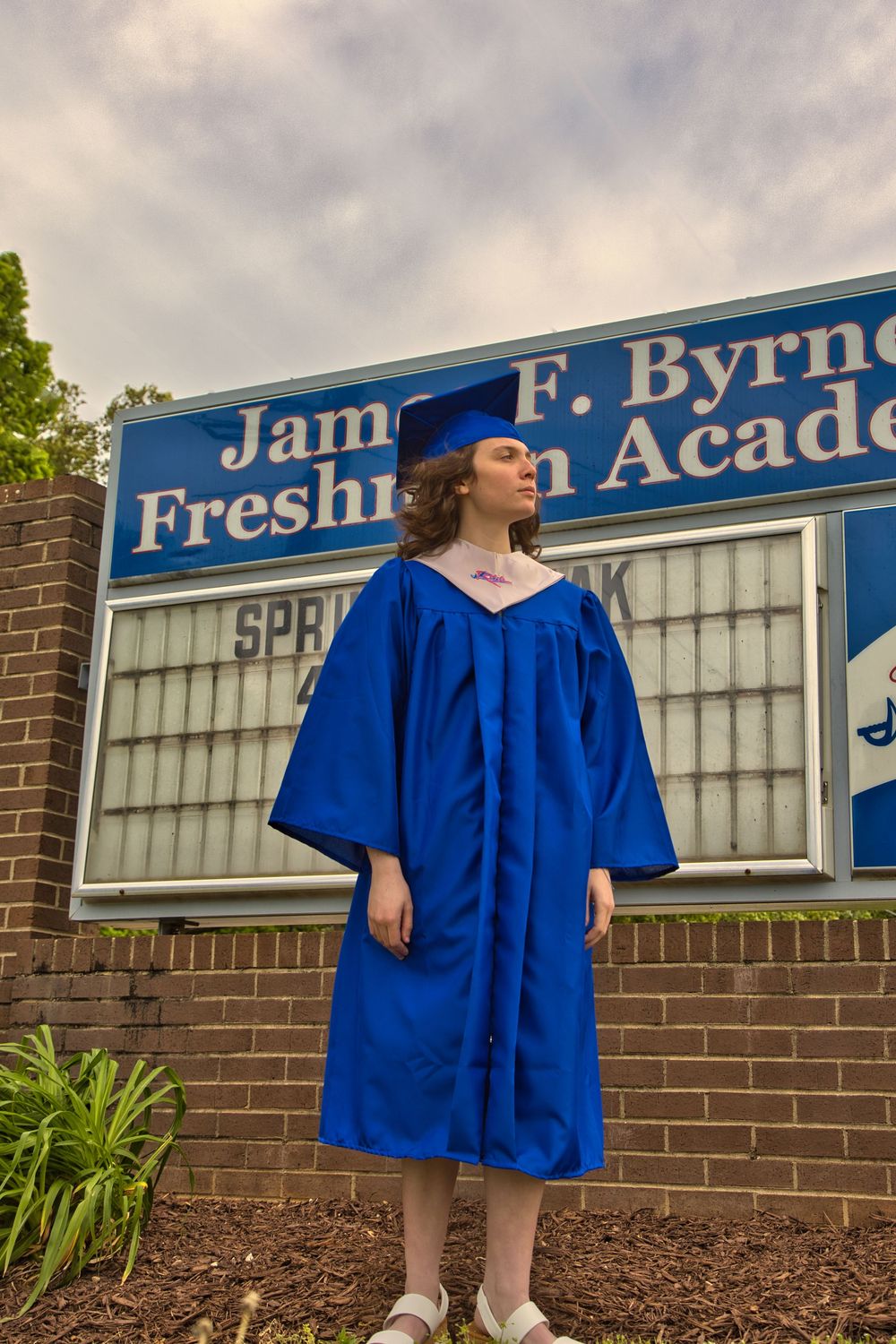 Graduate in blue cap and gown standing before James F. Byrnes Freshman Academy sign, in Duncan SC.