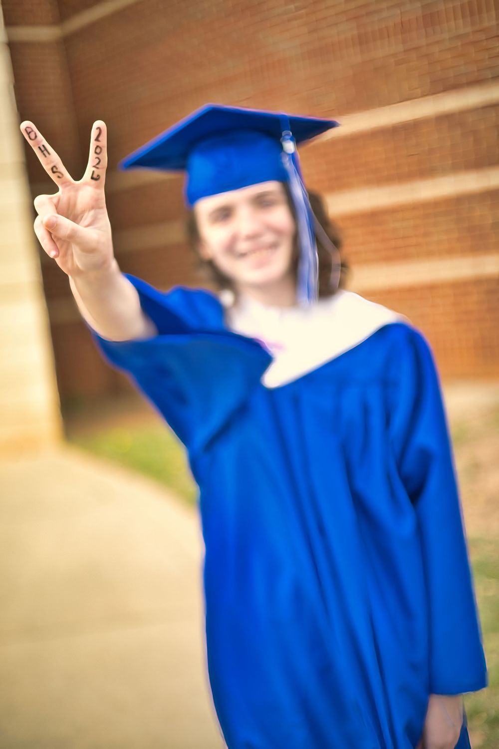 Graduate in blue cap and gown flashing peace sign with fingers toward camera.