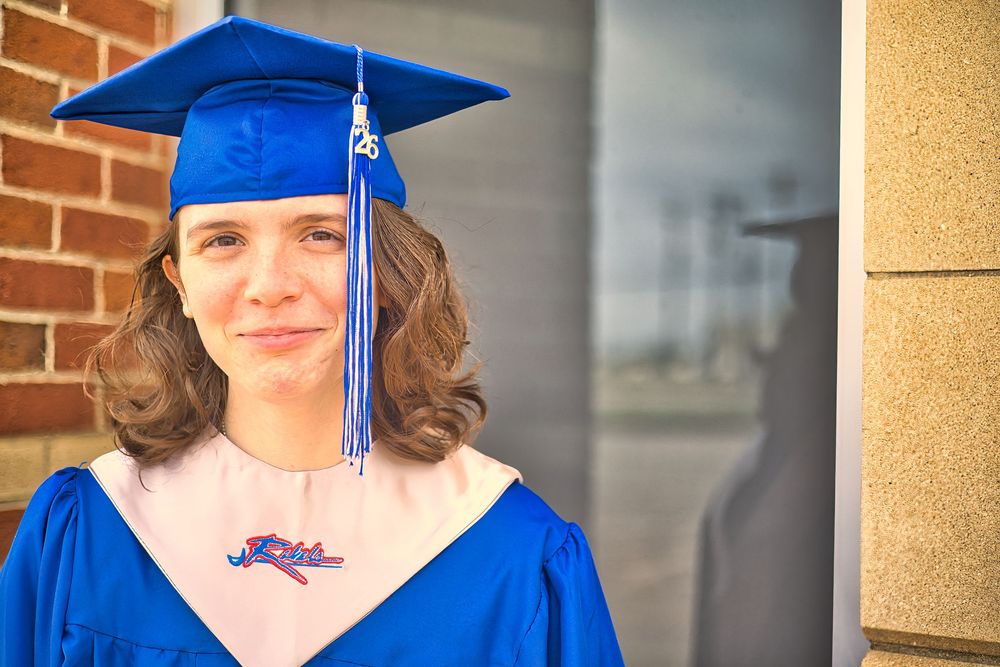 Young woman in blue graduation cap and gown standing near brick wall smiling.