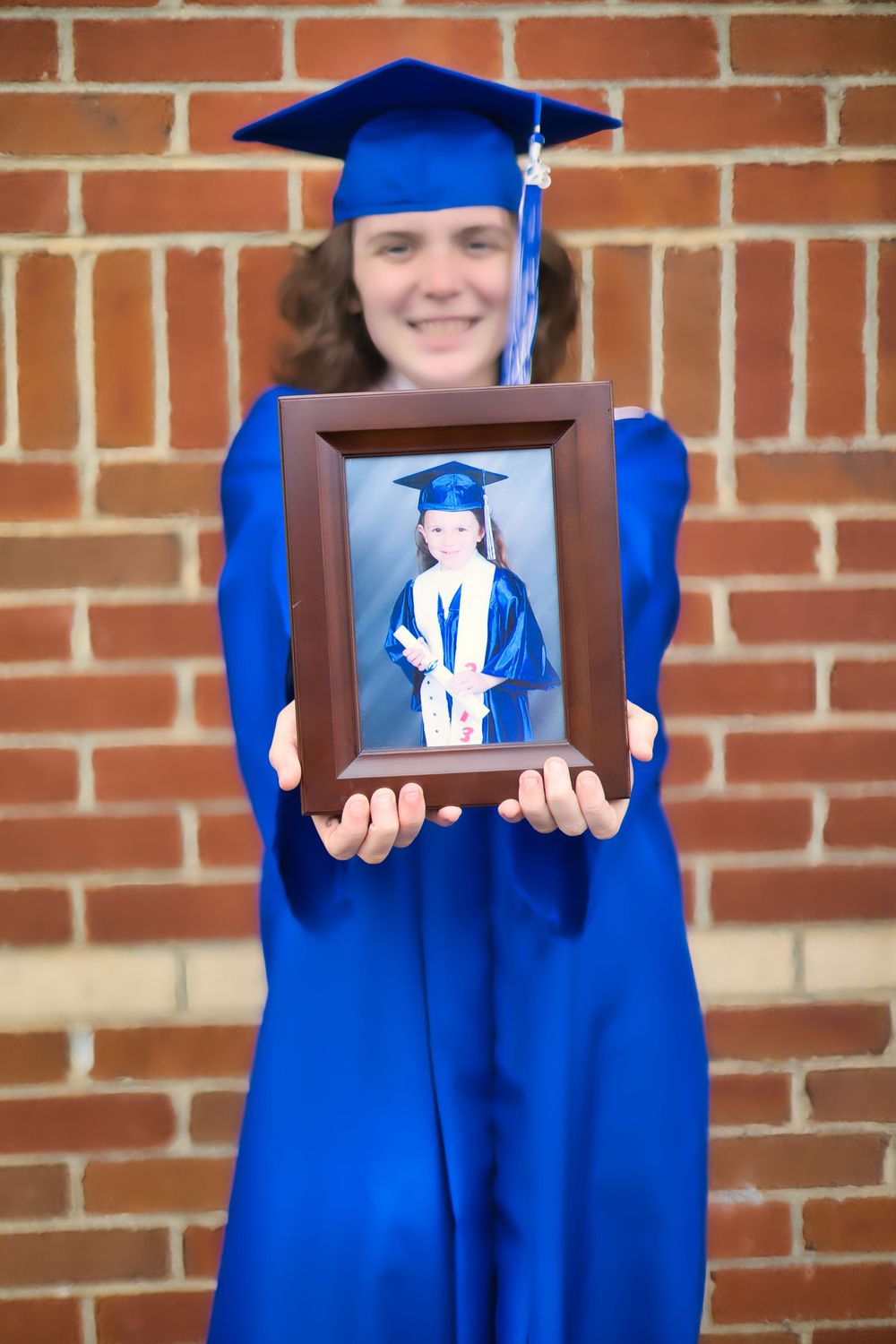 Graduate in blue cap and gown holding framed childhood photo in front of brick wall.