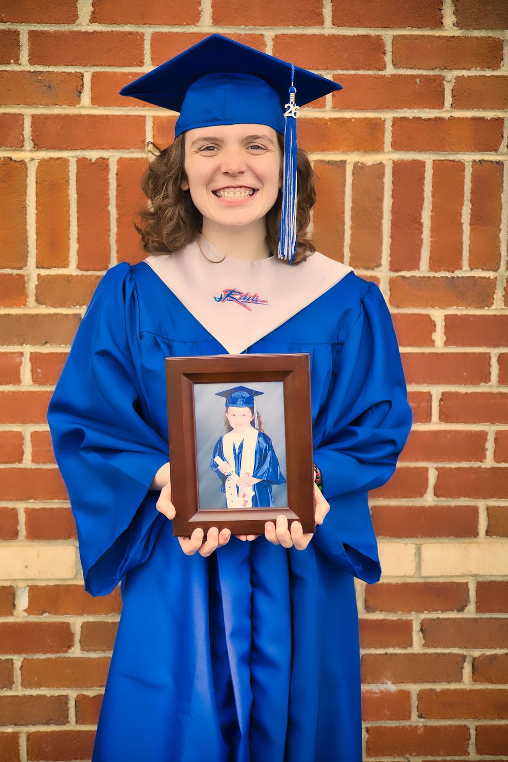 Smiling graduate in blue cap and gown holding framed childhood graduation photo against brick wall.