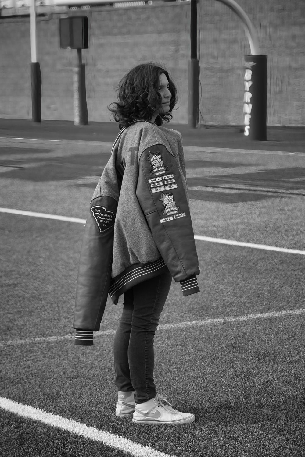 Black and white photo of student in varsity jacket standing on football field turf.