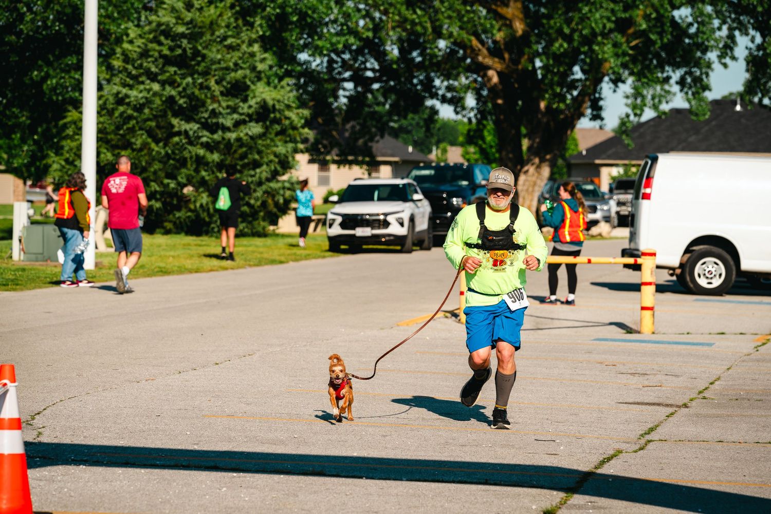 Runner in yellow shirt jogging with small dog on leash during outdoor road race event on sunny day.