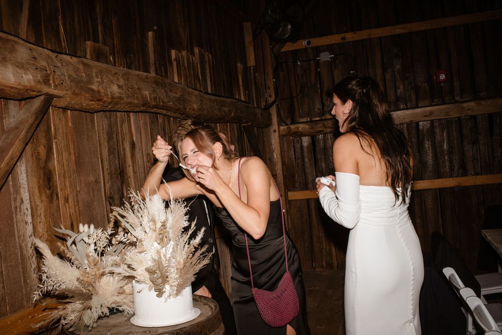 Bride and guests laughing together near a white wedding cake with pampas grass decor inside a rustic barn venue.