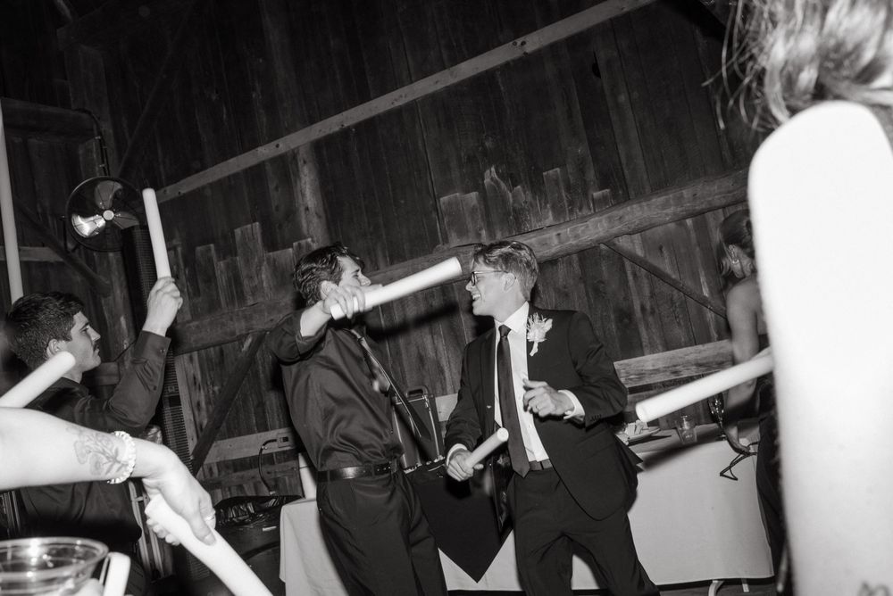 Black and white photo of men in suits dancing and celebrating at a rustic barn wedding reception.