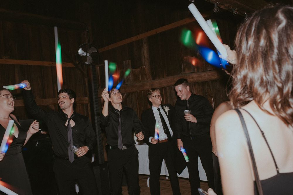 Group of men in black suits waving colorful glow sticks at a lively wedding reception dance floor.