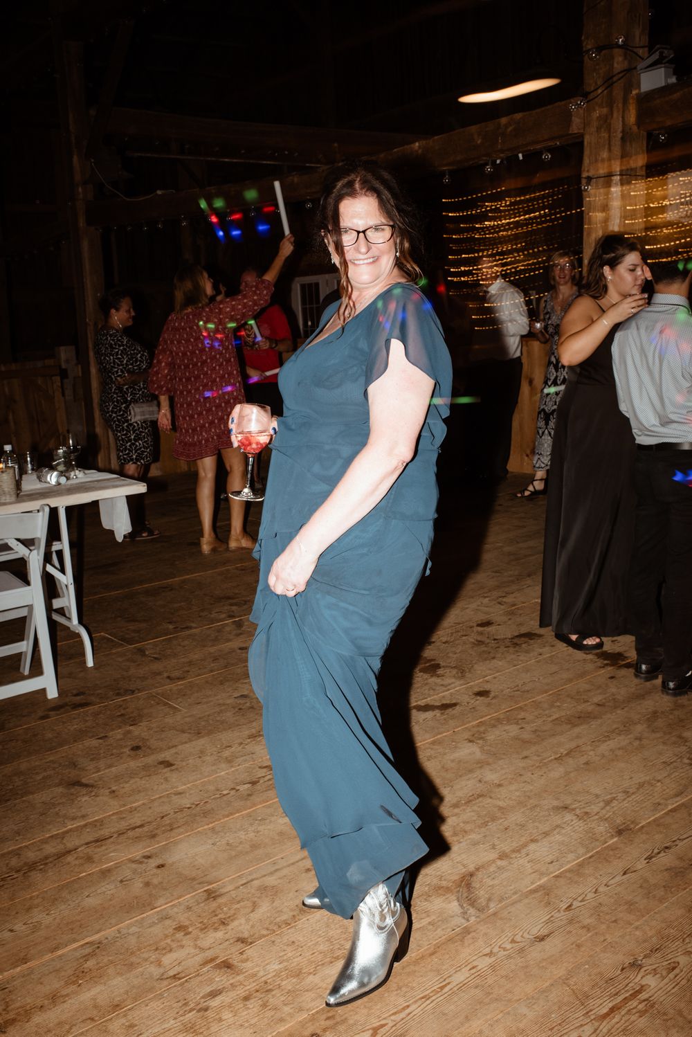 Woman in teal gown and silver boots dancing at a rustic barn wedding reception.