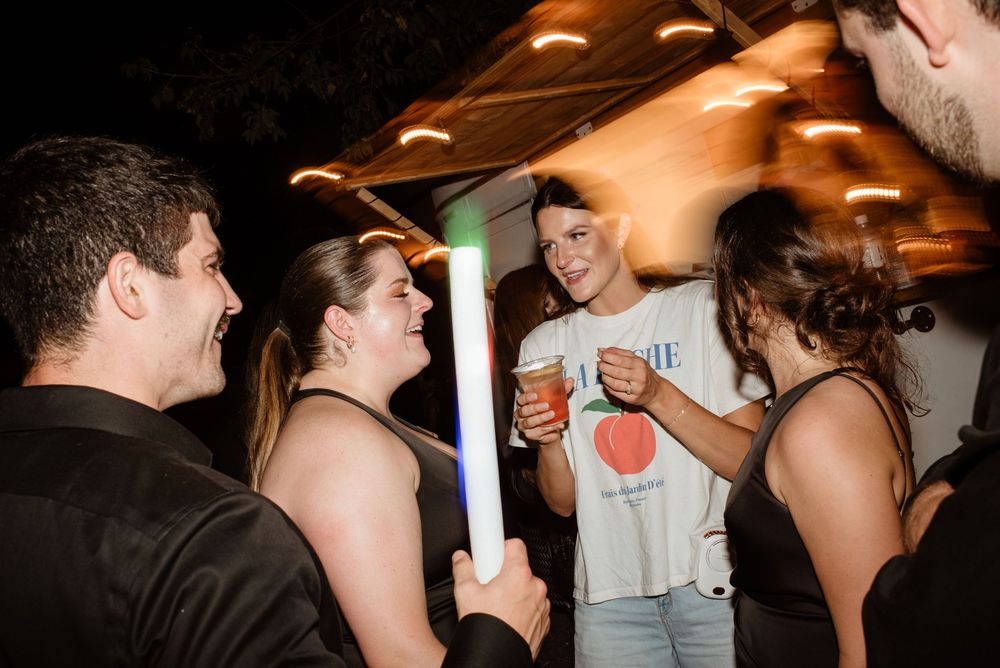 Group of young adults socializing at an outdoor evening party with string lights and colorful glow sticks.