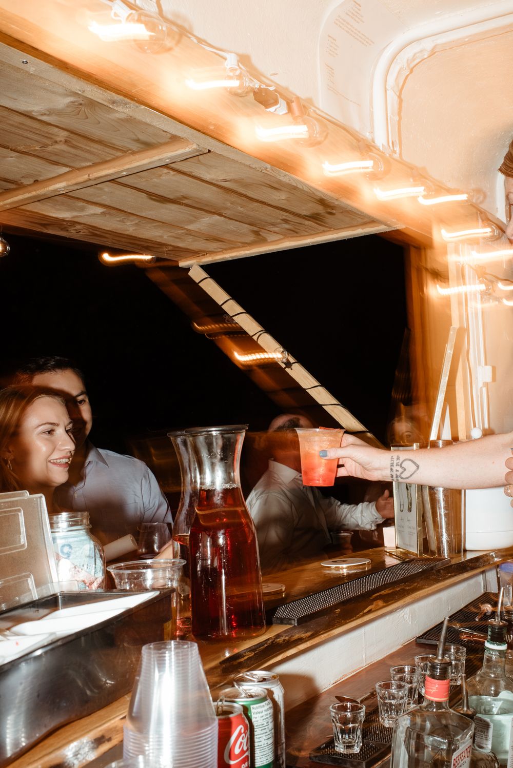 Bartender serving a pink cocktail at a warmly lit wooden bar with drinks and glassware on the counter.