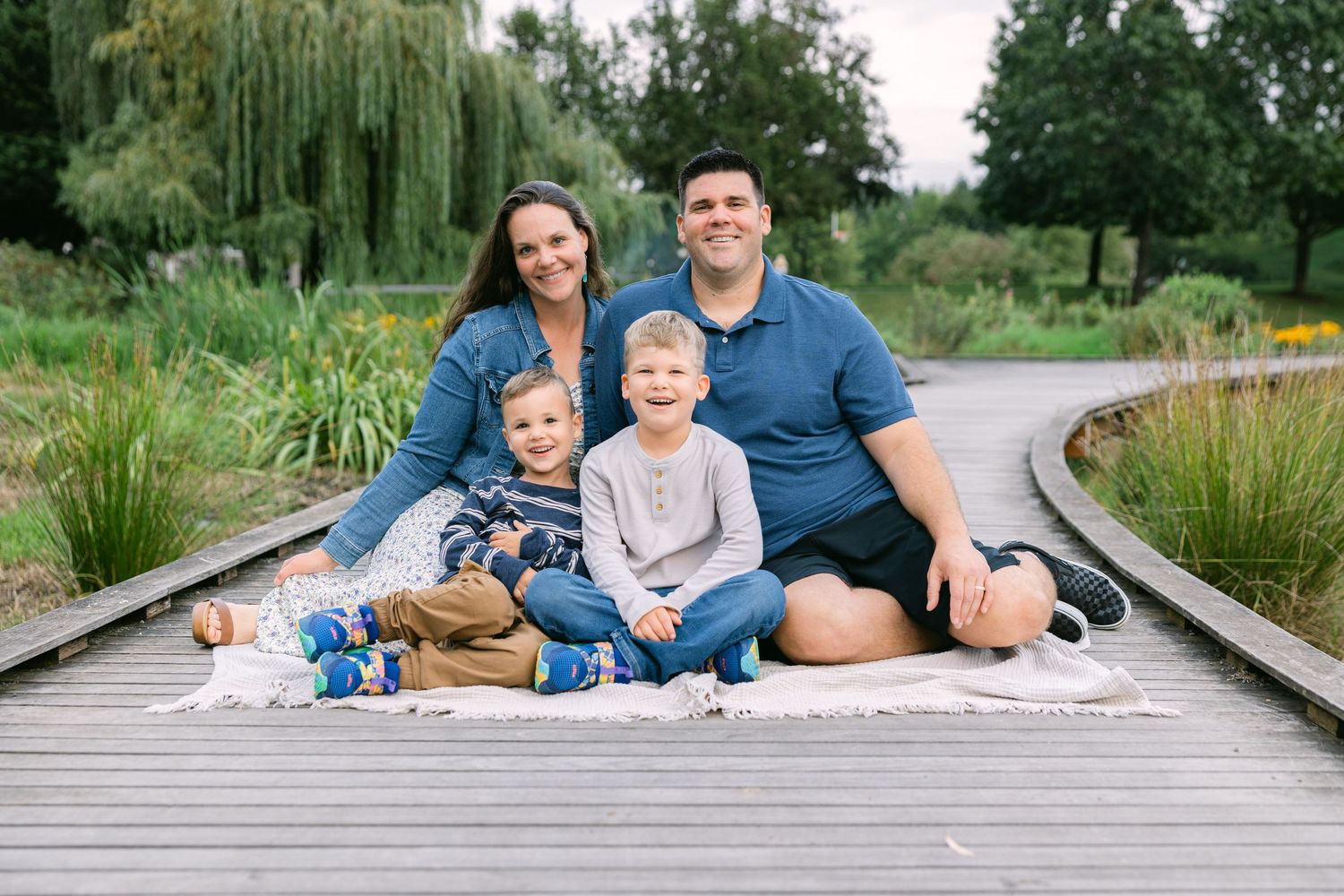 Family of four smiling on a wooden boardwalk surrounded by lush greenery, with two young boys seated in front.