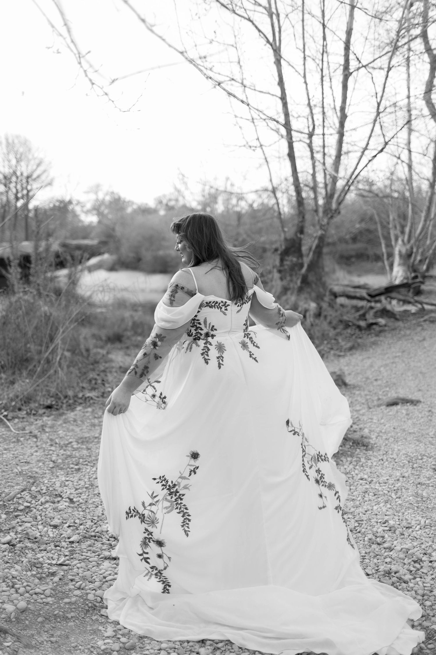 Black and white photo of a bride in a floral embroidered gown holding her skirt outdoors.